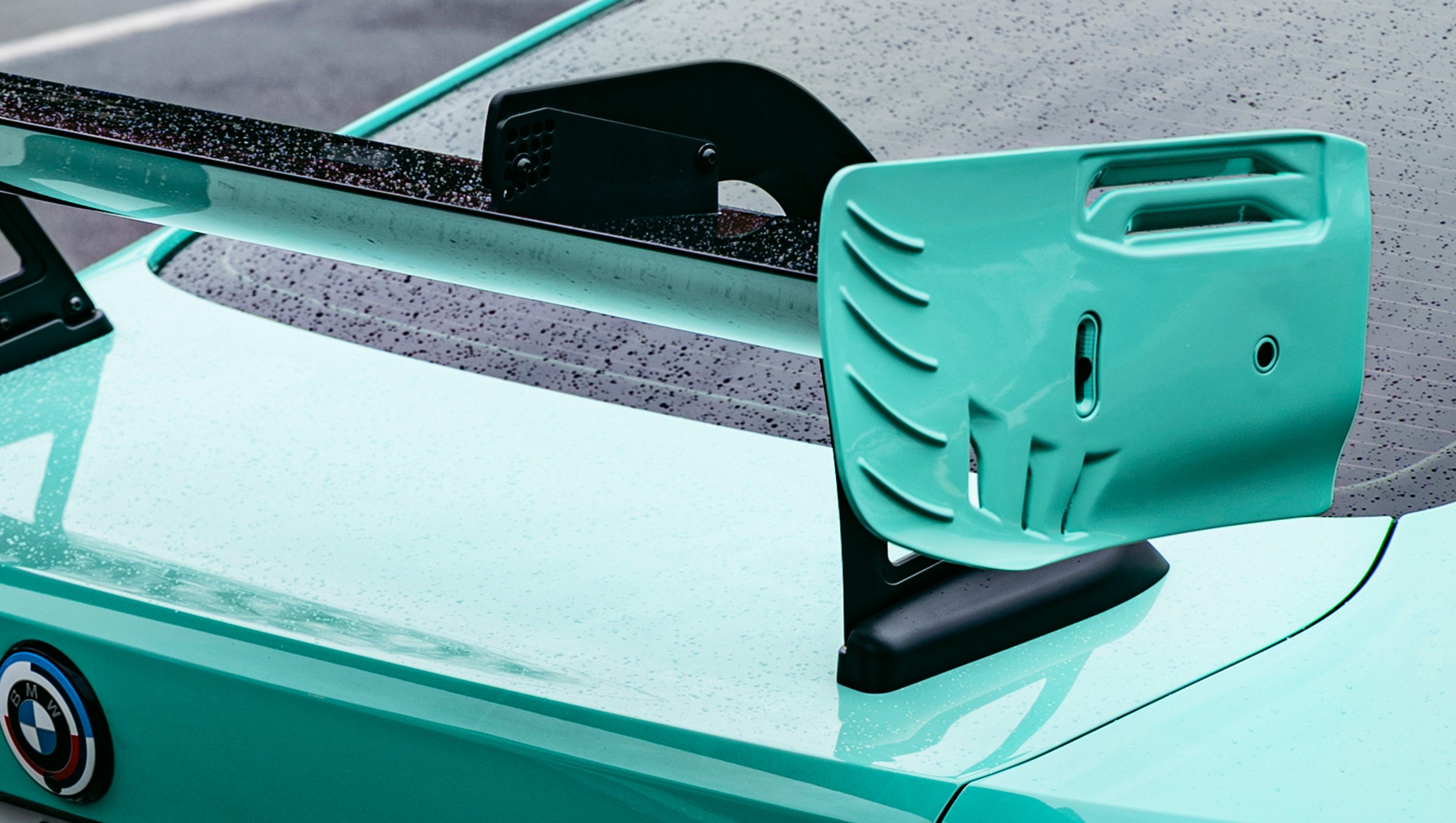 Mint green BMW G82 M4 with carbon fiber front splitter, side skirts, and large rear wing parked on a wet urban street outside a modern building.