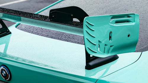 Mint green BMW G82 M4 with carbon fiber front splitter, side skirts, and large rear wing parked on a wet urban street outside a modern building.