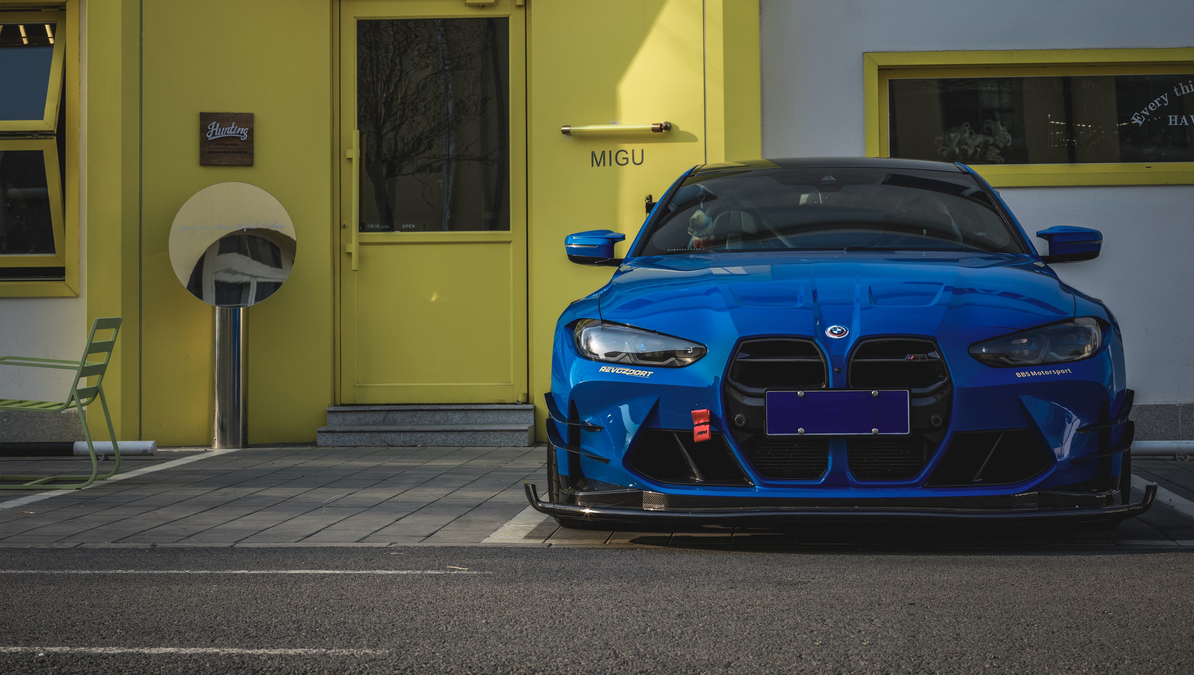 Blue BMW G82 M4 with carbon fiber front splitter, side skirts, rear wing, and aftermarket wheels parked in a modern industrial courtyard with colorful architectural walls.