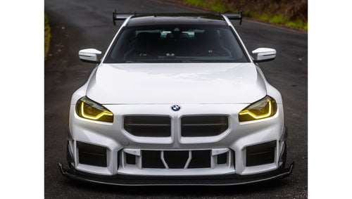 White widebody BMW G82 M4 with aggressive front bumper, canards, deep front splitter, large rear wing, and yellow LED headlights parked on a wet road surrounded by dense tropical forest.