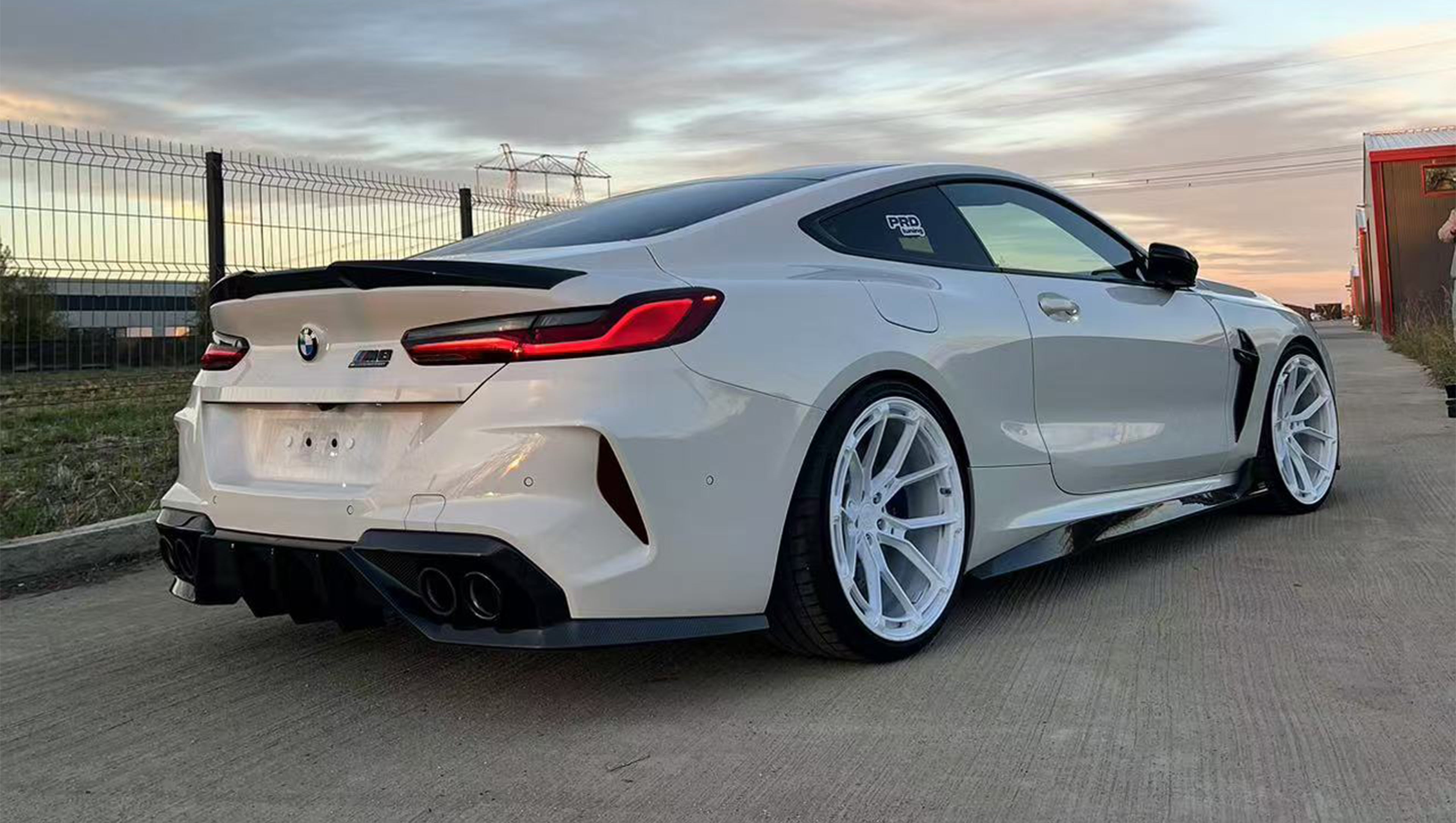 White BMW F92 M8 featuring a REVOZPORT carbon fiber front lip, side skirts, black front grille, and white multi-spoke forged wheels parked at sunset on an open road.