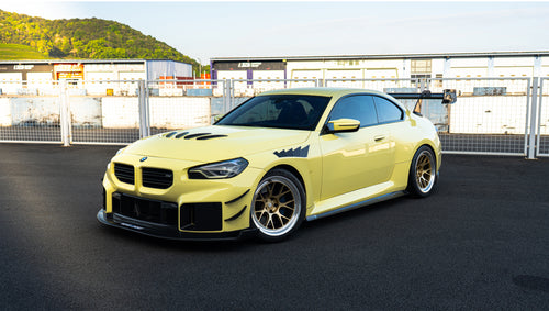 Pastel yellow BMW G82 M4 with carbon fiber front splitter, hood vents, side canards, bronze wheels, and large rear wing parked at a race track facility during golden hour.