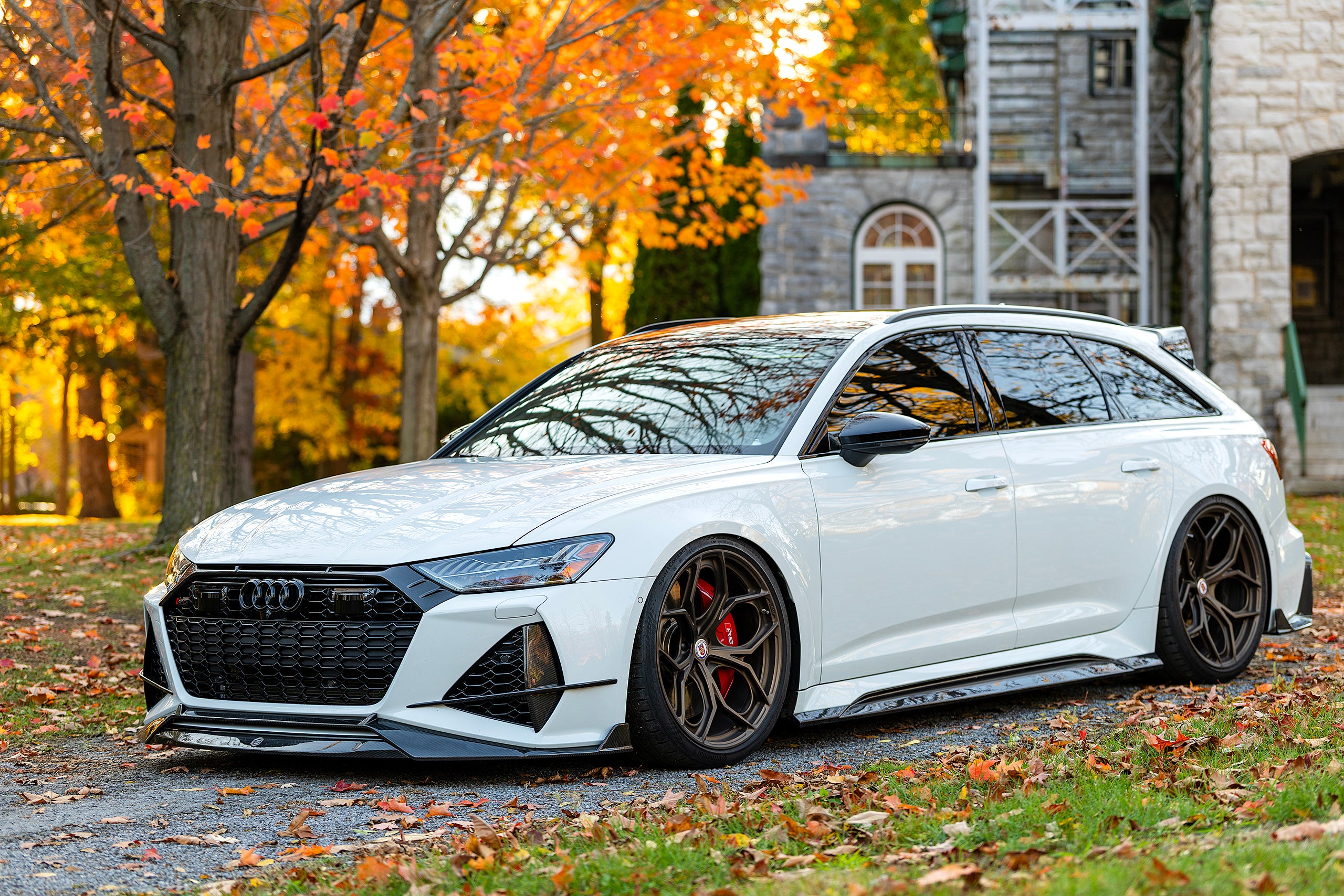White Audi RS6 Avant featuring a REVOZPORT carbon fiber body kit with front lip, side skirts, and aggressive aero styling parked in front of a historic stone mansion during autumn.