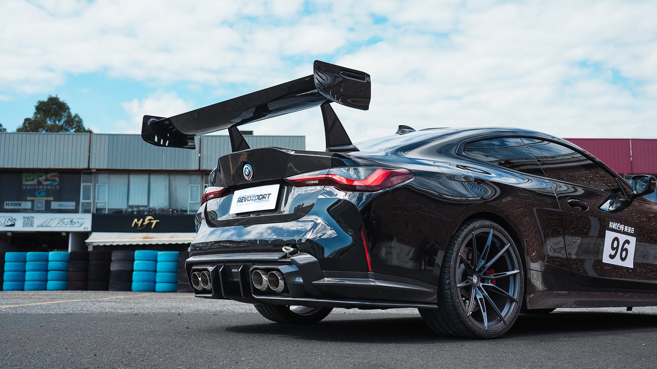 Black BMW G82 M4 track build with carbon fiber vented fenders, side skirts, rear diffuser, and oversized GT wing parked at a professional racing circuit pit lane.