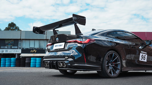 Black BMW G82 M4 track build with carbon fiber vented fenders, side skirts, rear diffuser, and oversized GT wing parked at a professional racing circuit pit lane.