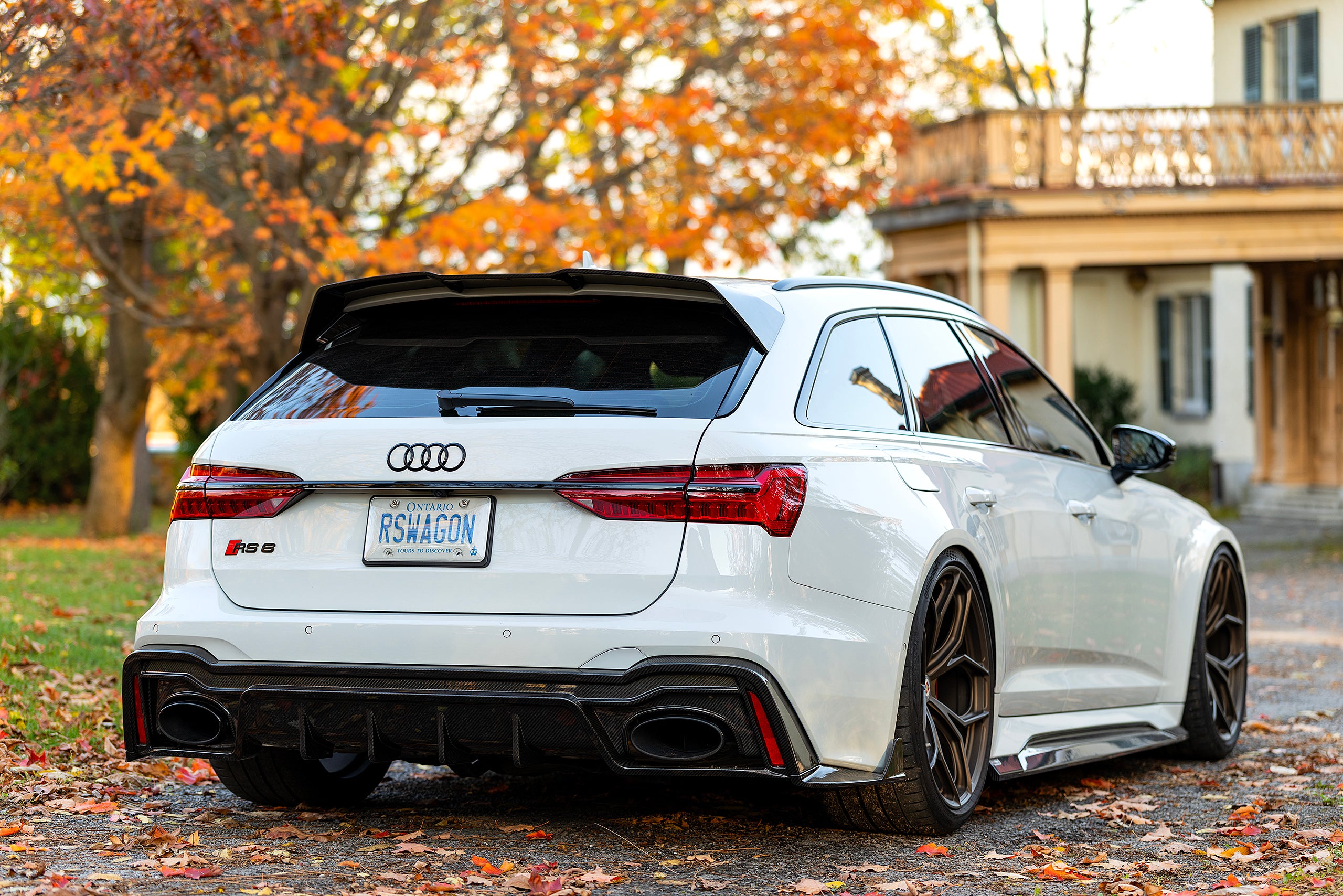 White Audi RS6 Avant featuring a REVOZPORT carbon fiber body kit with front lip, side skirts, and aggressive aero styling parked in front of a historic stone mansion during autumn.