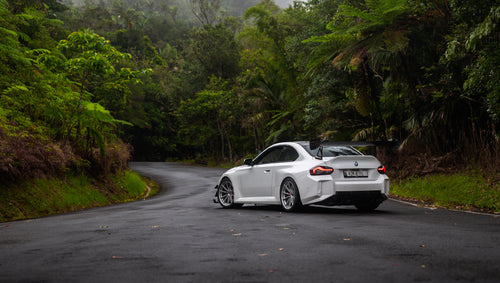 White widebody BMW G82 M4 with aggressive front bumper, canards, deep front splitter, large rear wing, and yellow LED headlights parked on a wet road surrounded by dense tropical forest.