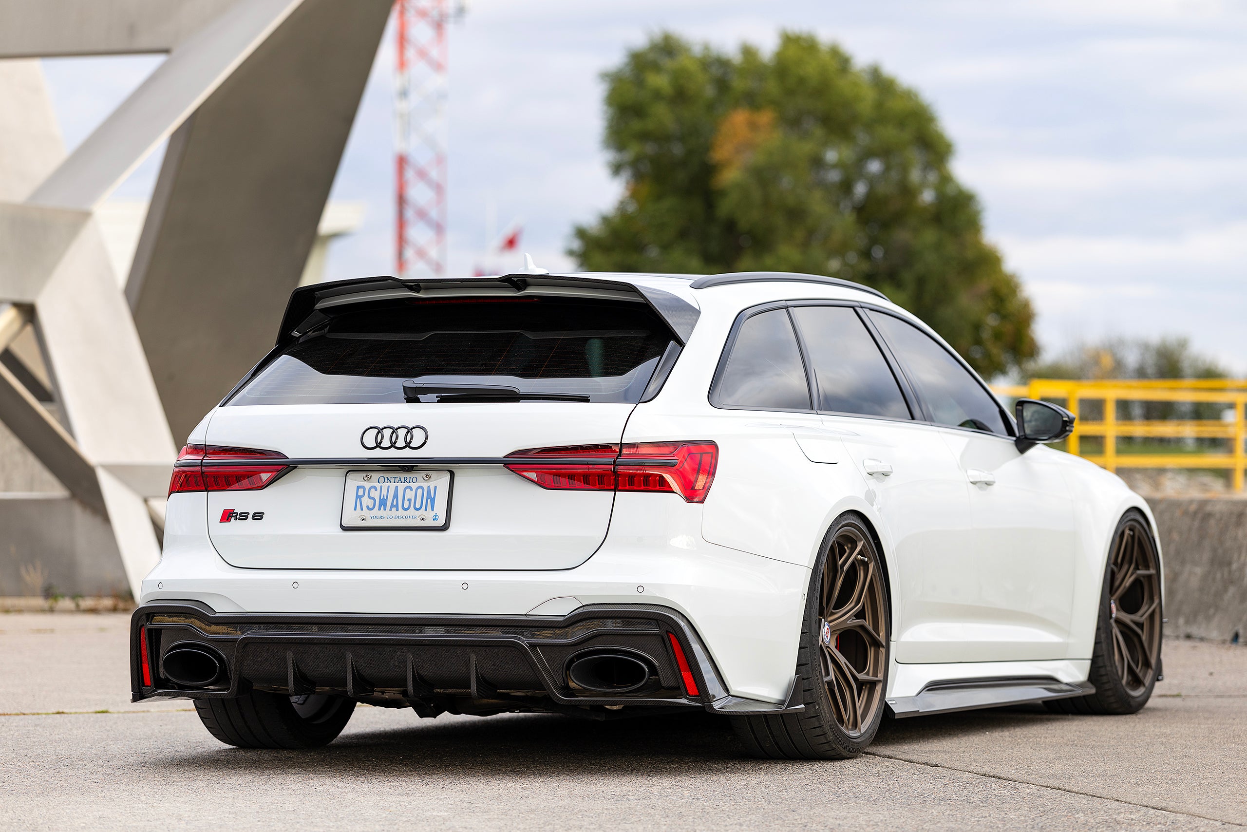 White Audi RS6 Avant featuring a REVOZPORT carbon fiber body kit with front lip, side skirts, rear diffuser, and lowered stance parked in a modern urban waterfront setting.