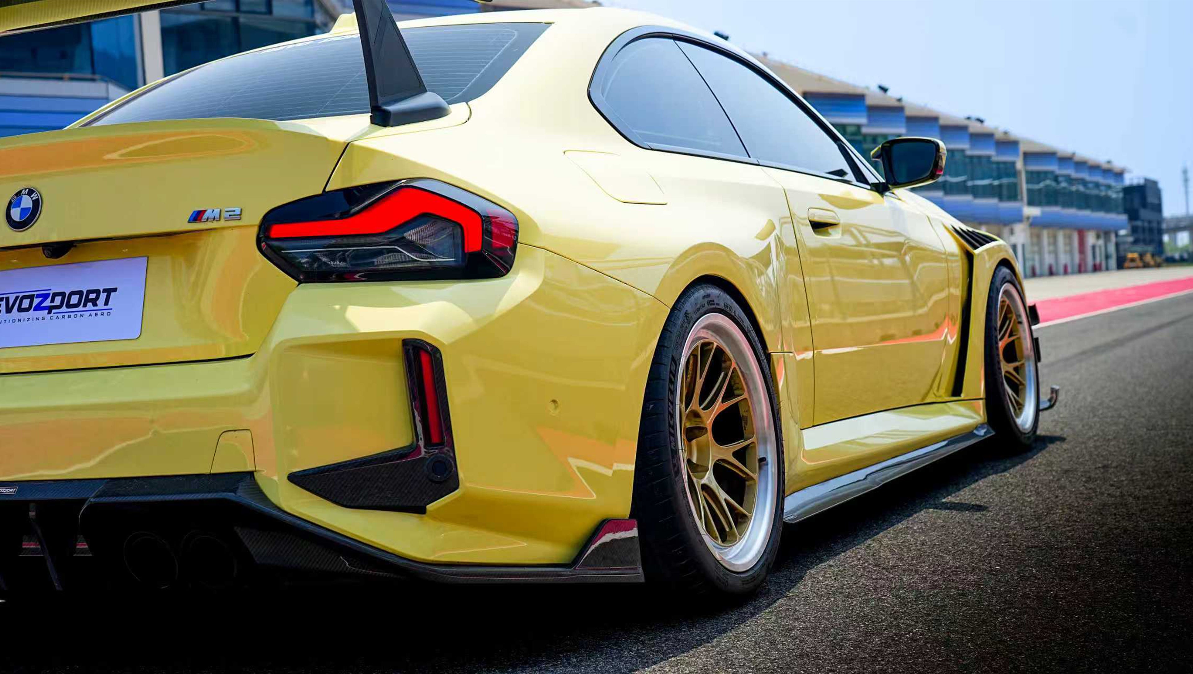 Yellow BMW G82 M4 with aggressive carbon fiber front splitter, vented hood, side canards, and bronze wheels parked on a race track pit lane.