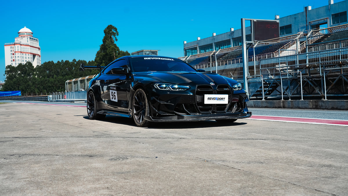Black BMW G82 M4 track build with carbon fiber vented fenders, side skirts, rear diffuser, and oversized GT wing parked at a professional racing circuit pit lane.