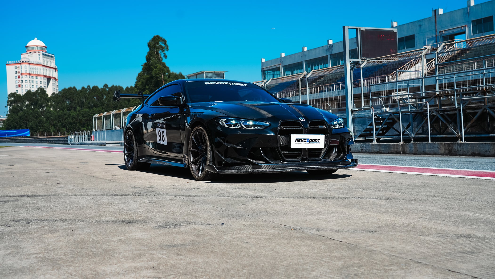 Black BMW G82 M4 track build with carbon fiber vented fenders, side skirts, rear diffuser, and oversized GT wing parked at a professional racing circuit pit lane.
