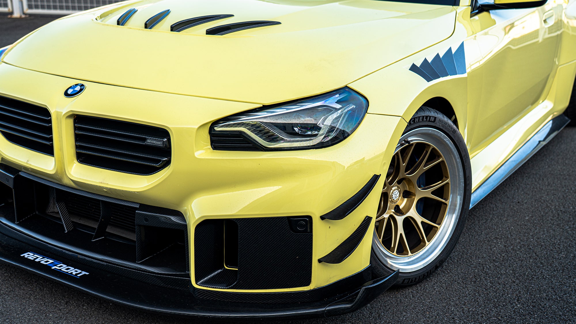 Close-up detail of a BMW front end featuring aggressive carbon fiber aero, vented hood, forged bronze wheels, and track-inspired styling.