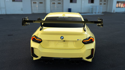 Pastel yellow BMW G82 M4 with carbon fiber front splitter, hood vents, side canards, bronze wheels, and large rear wing parked at a race track facility during golden hour.
