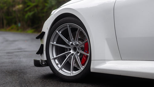 White widebody BMW G82 M4 with aggressive front bumper, canards, deep front splitter, large rear wing, and yellow LED headlights parked on a wet road surrounded by dense tropical forest.