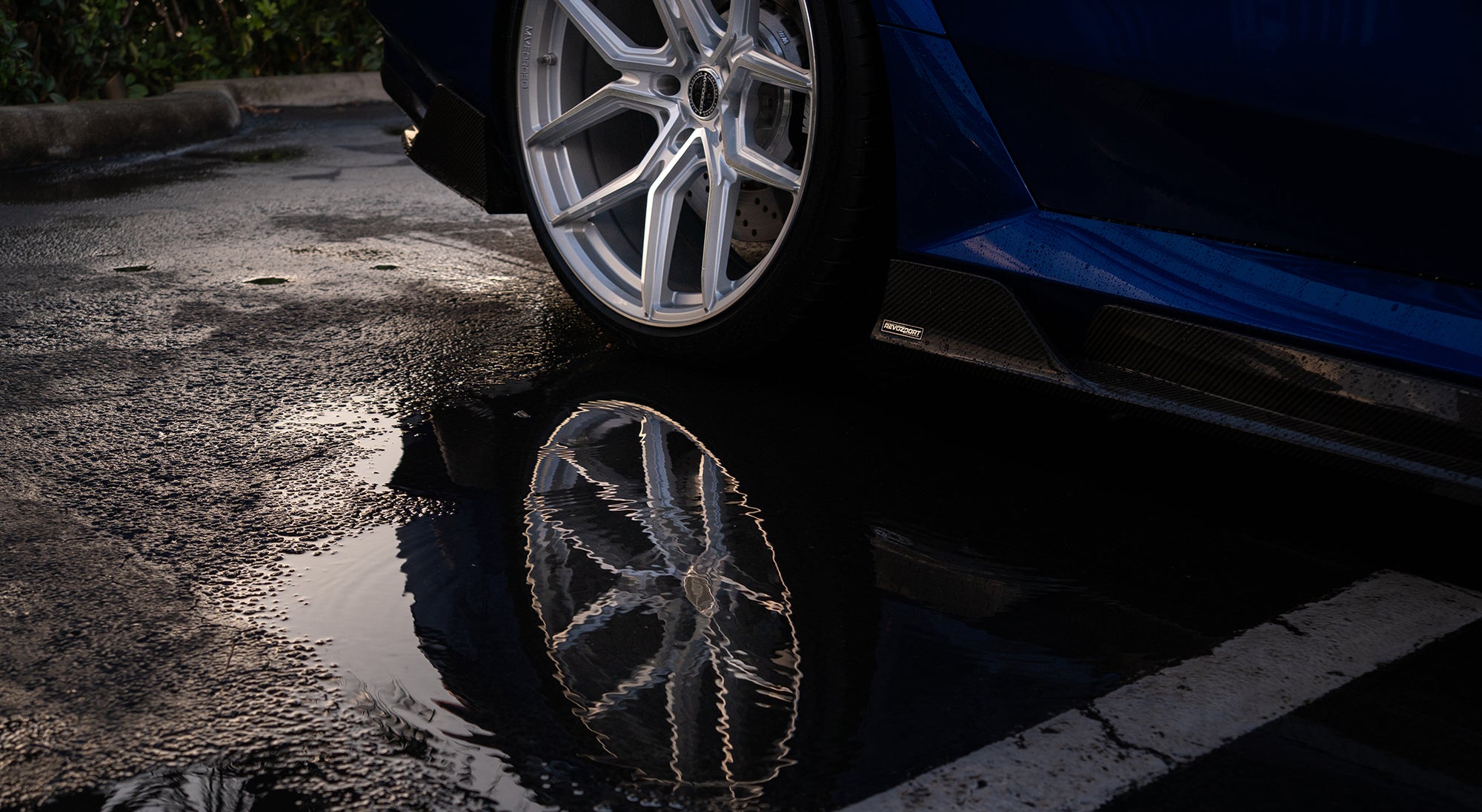 Blue BMW G90 M5 featuring a REVOZPORT carbon fiber front lip, side skirts, aggressive aero styling, and silver forged wheels parked on a wet road with palm trees at sunset.