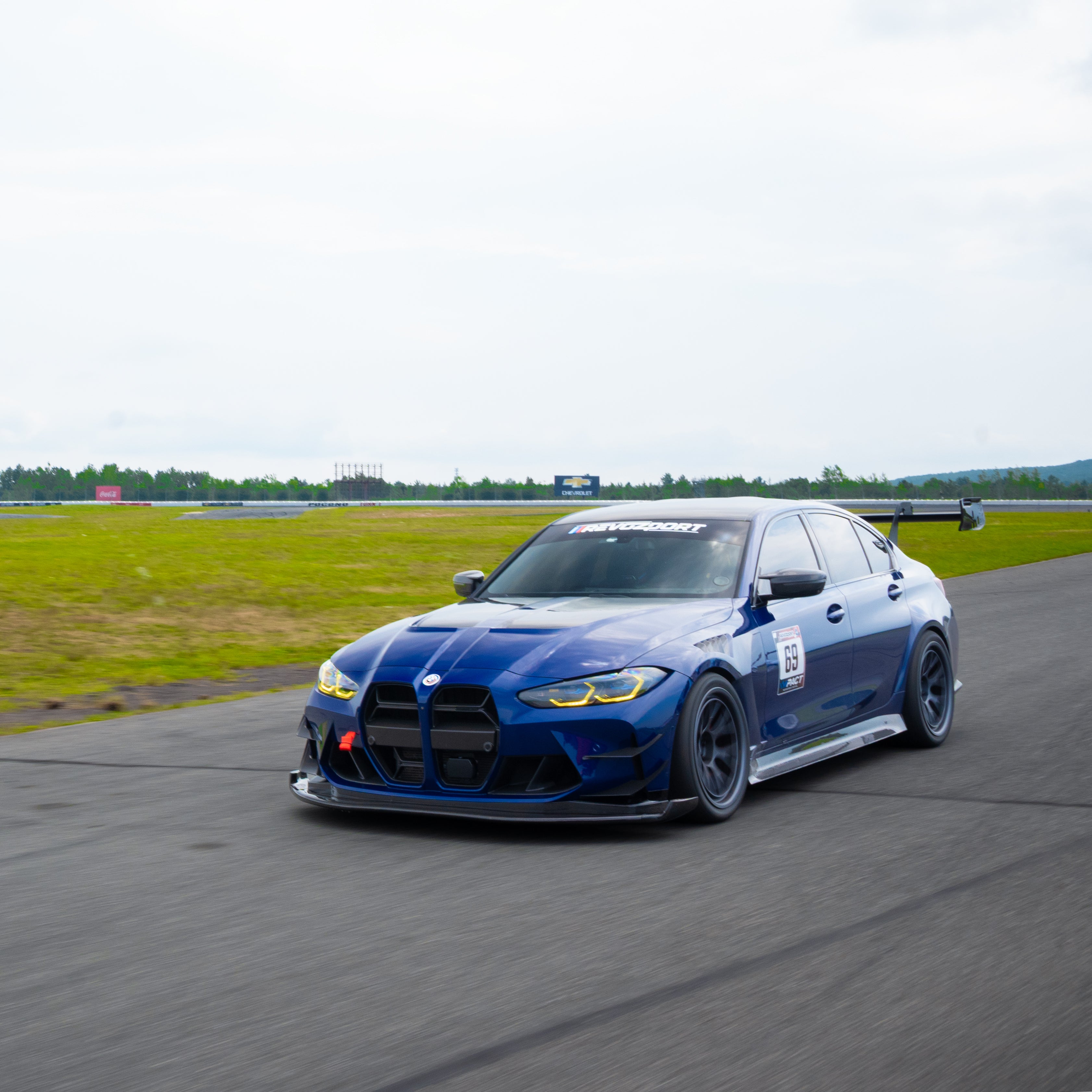 Blue BMW G80 M3 with aggressive carbon fiber aero kit, front splitter, vented fenders, and side skirts parked beside a professional race track during sunset.