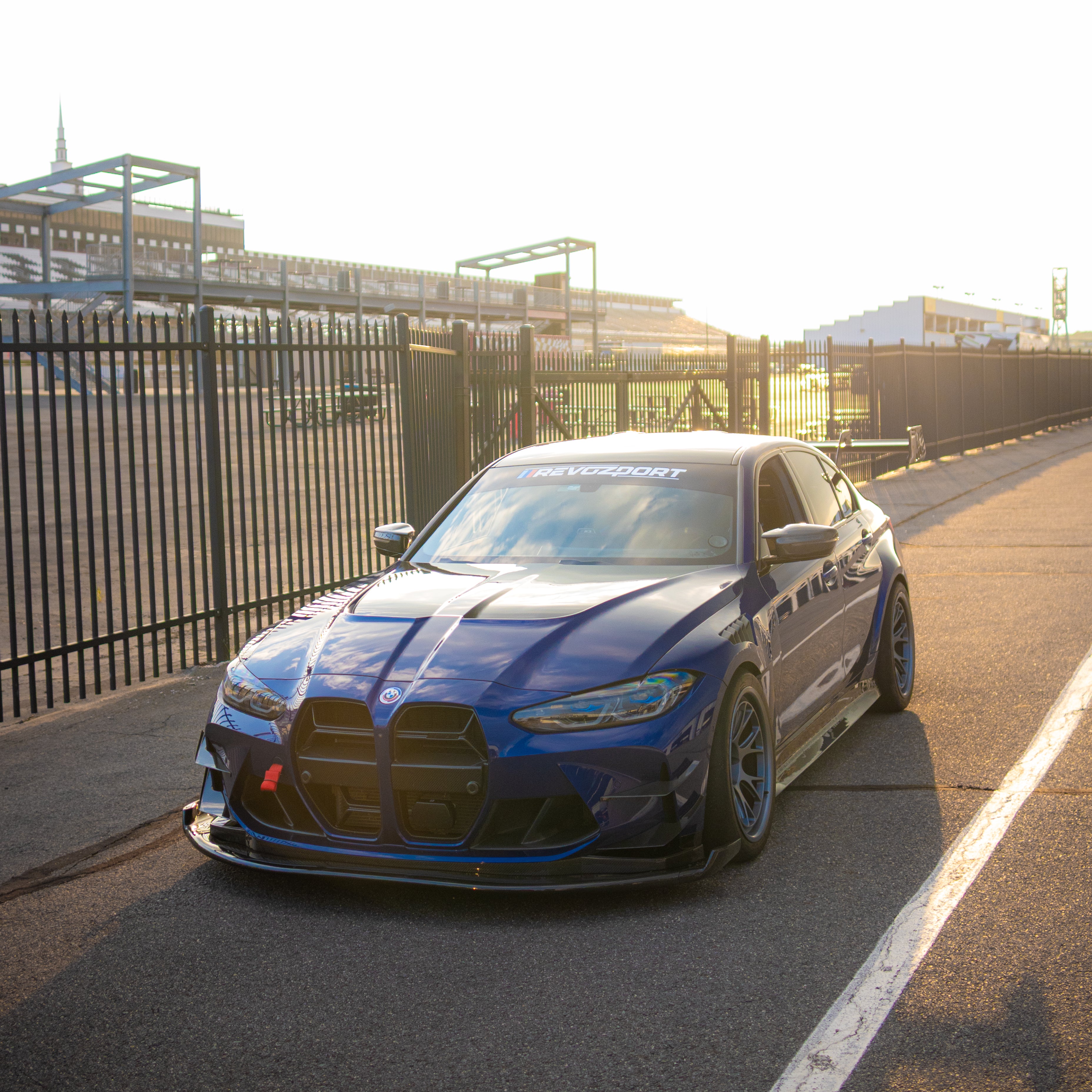 Blue BMW G80 M3 with aggressive carbon fiber aero kit, front splitter, vented fenders, and side skirts parked beside a professional race track during sunset.