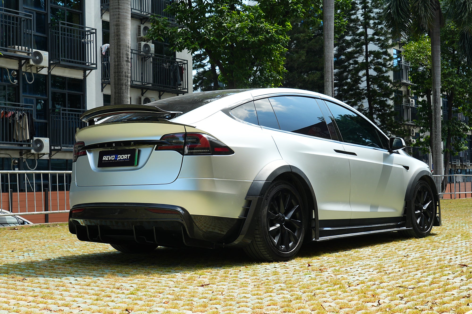 Silver Tesla Model X featuring a REVOZPORT body kit with carbon fiber front lip, side skirts, wide fender flares, and lowered stance parked outdoors in sunlight.