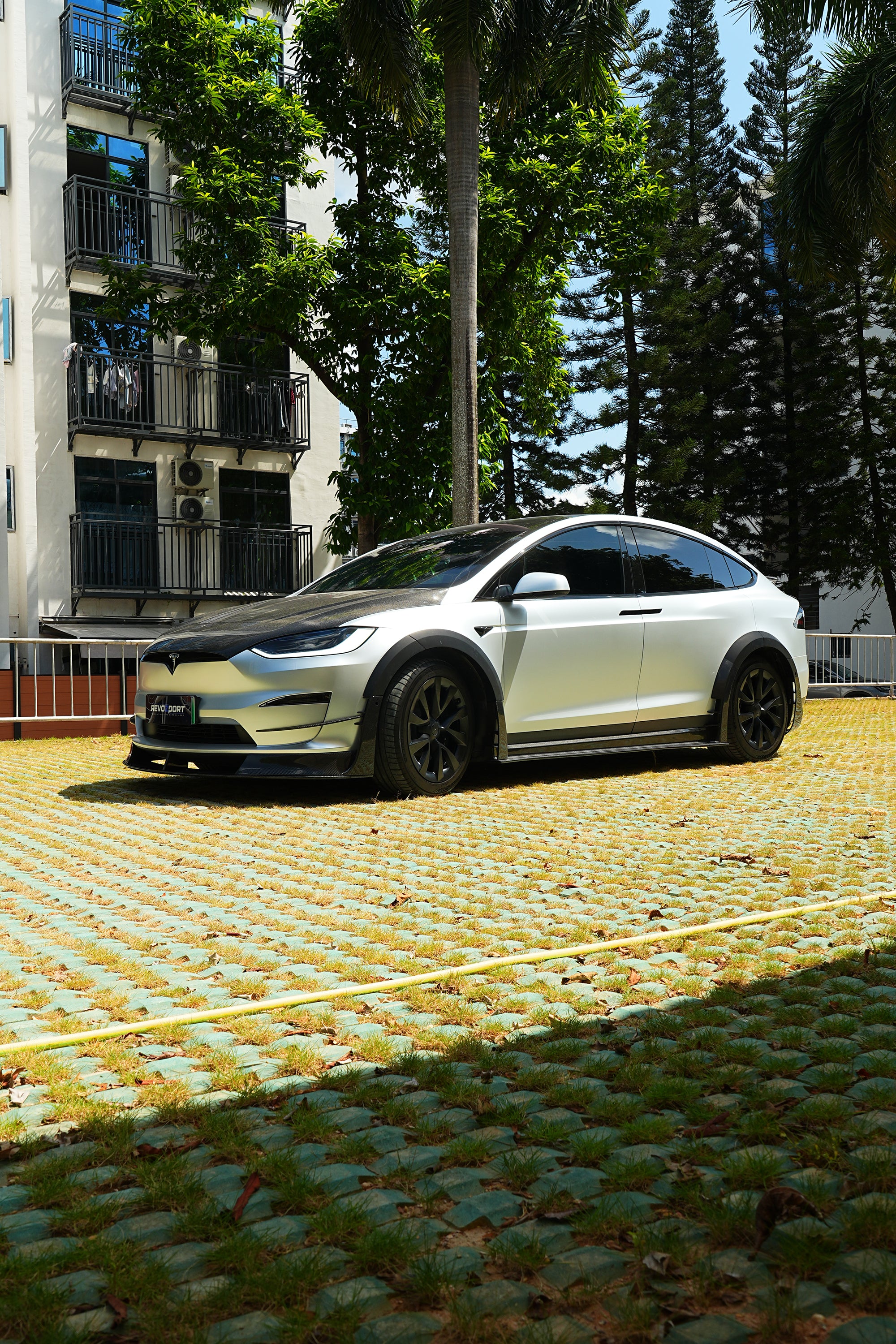 Silver Tesla Model X featuring a REVOZPORT body kit with carbon fiber front lip, side skirts, wide fender flares, and lowered stance parked outdoors in sunlight.