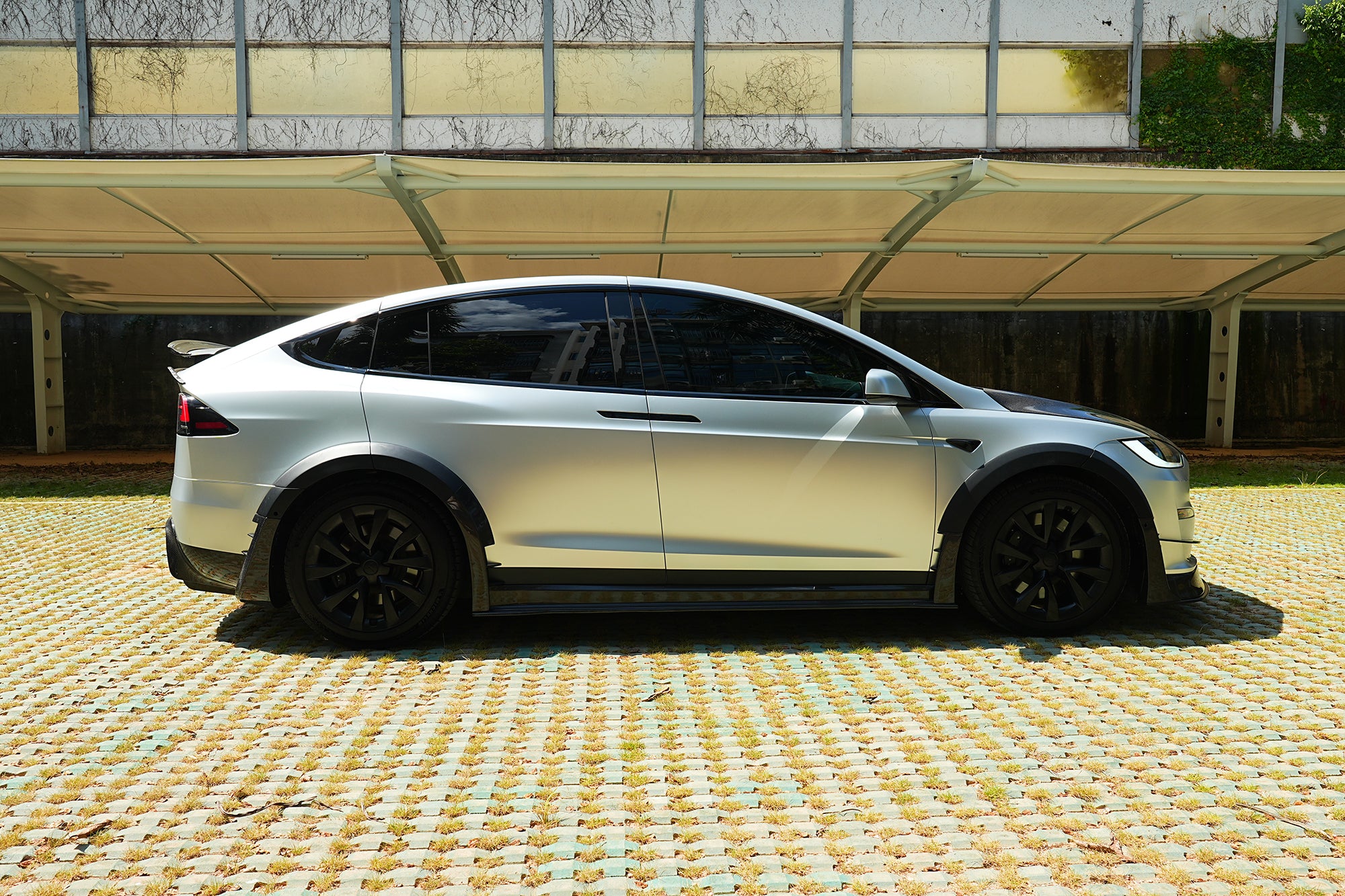 Silver Tesla Model X featuring a REVOZPORT body kit with carbon fiber front lip, side skirts, wide fender flares, and lowered stance parked outdoors in sunlight.