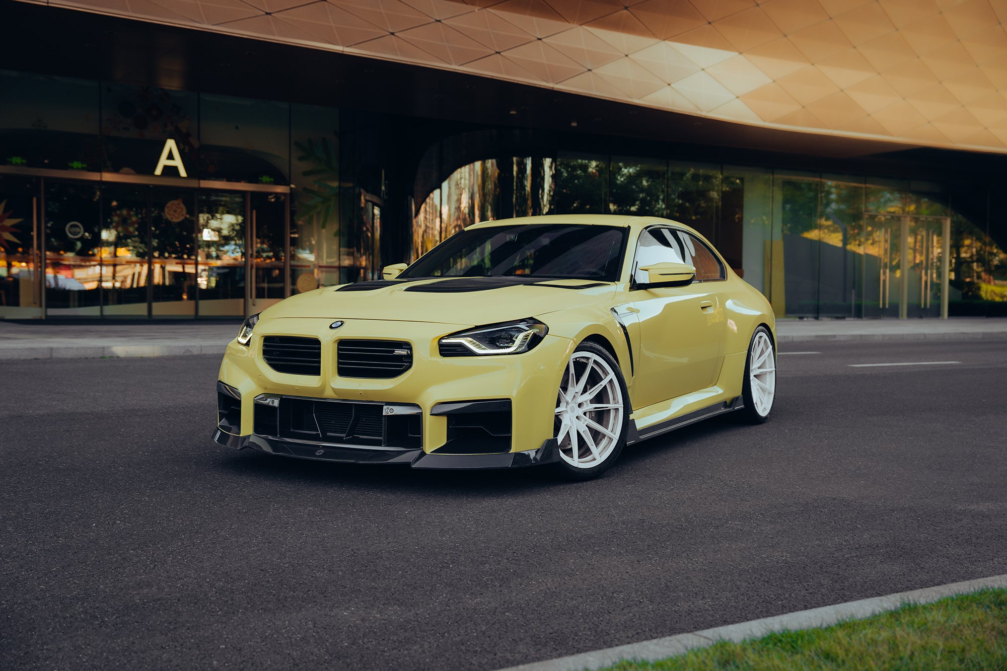 Yellow BMW with carbon fiber body kit parked outside a modern building, highlighting front lip, side skirts, hood vents, and white wheels.