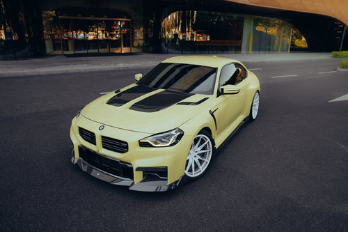 Yellow BMW with carbon fiber body kit parked outside a modern building, highlighting hood vents, front lip, and white wheels.