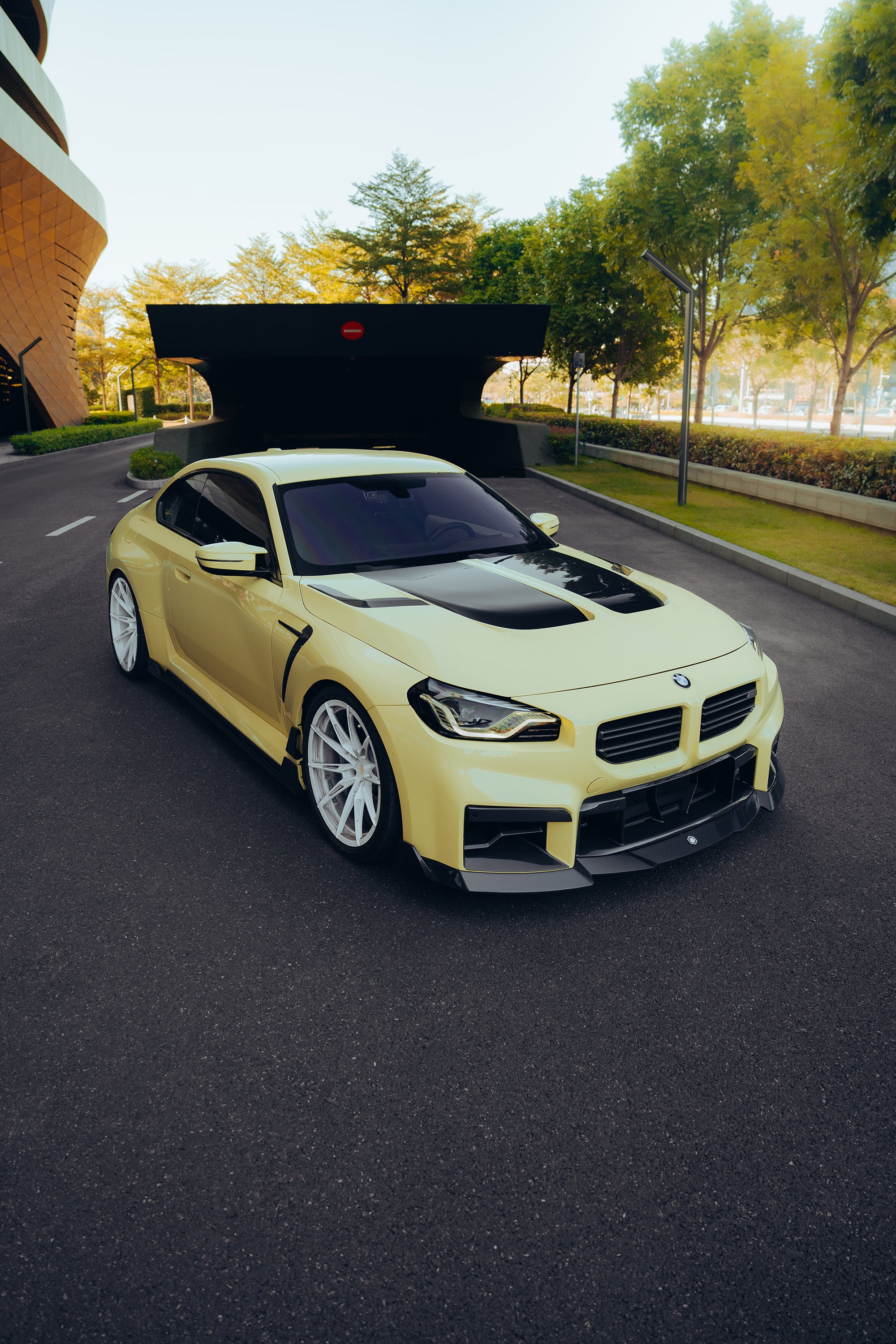 Yellow BMW with carbon fiber body kit on a modern street, highlighting hood vents, front lip, and white wheels.