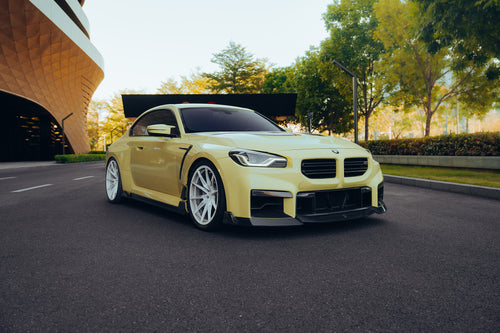 Yellow BMW with carbon fiber body kit parked on a modern street, highlighting front lip, side skirts, and white wheels.