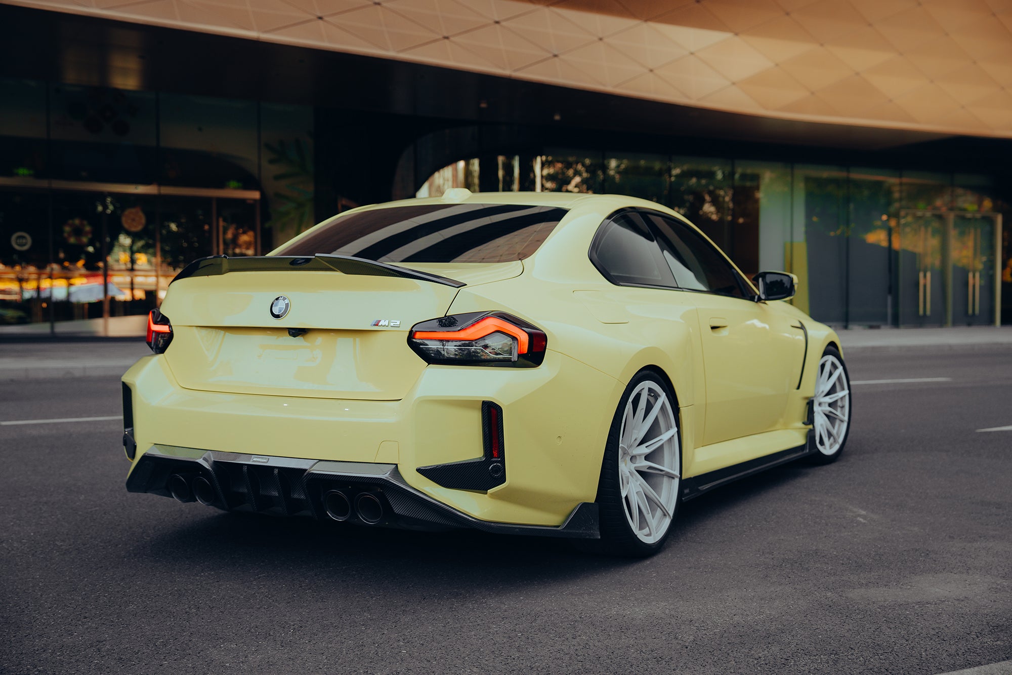 Yellow BMW with carbon fiber rear spoiler, diffuser, and side skirts parked outside a modern building, emphasizing rear aero styling.