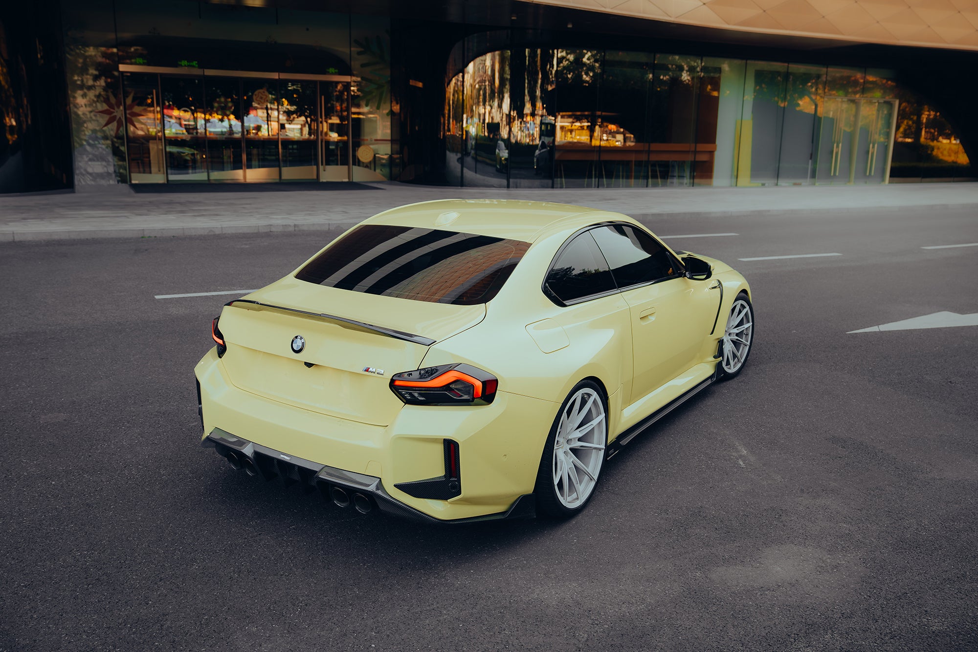 Yellow BMW with carbon fiber rear spoiler, diffuser, and side skirts parked outside a modern building, emphasizing sleek rear styling and stance.