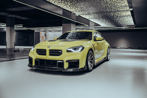 The BMW M2, wrapped in carbon fiber, sits in a high-end studio with lights on, equipped with high-end wheels, side skirts, and front lip
