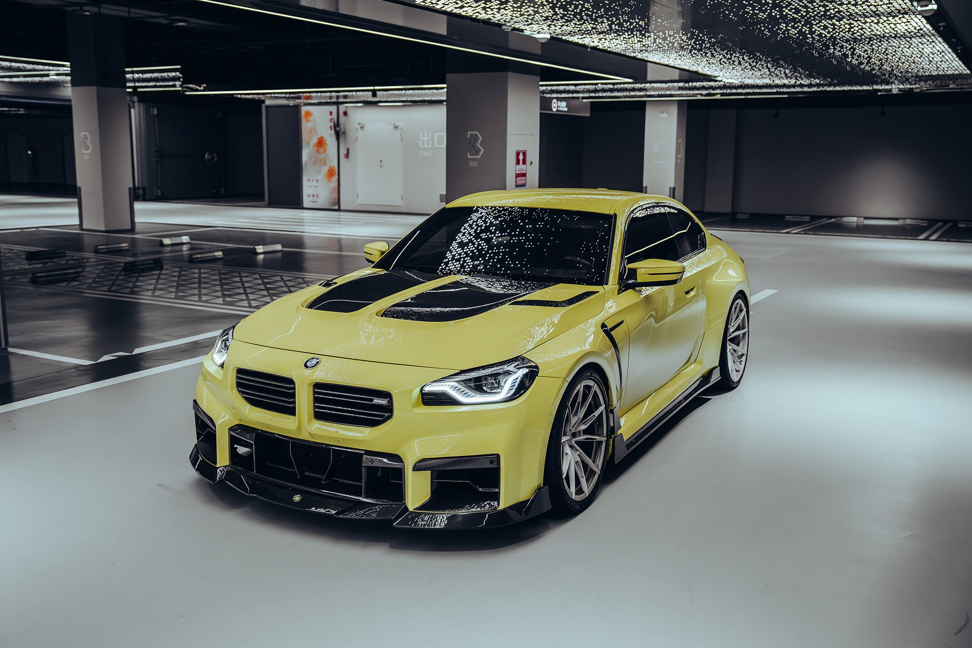 Yellow BMW with carbon fiber body kit parked in a modern garage, highlighting front lip, hood vents, and side skirt styling.