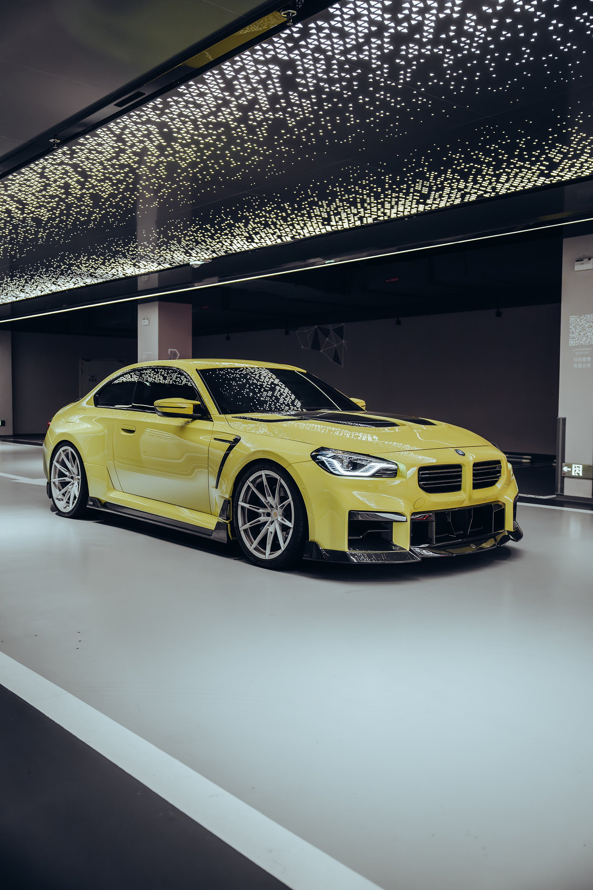 The BMW M2, wrapped in carbon fiber, sits in a high-end studio with lights on, equipped with high-end wheels, side skirts, and front lip