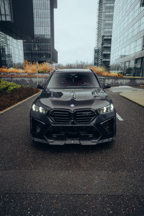 Black BMW X5M featuring a REVOZPORT carbon fiber body kit with aggressive front lip, side skirts, enlarged front air intakes, and sporty aero styling parked in a modern urban business district.