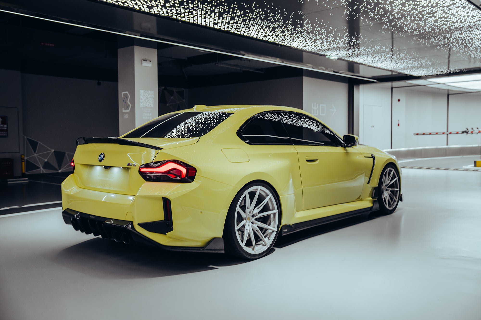 Yellow BMW with carbon fiber body kit parked in a modern garage, highlighting rear spoiler, side skirts, and diffuser styling.