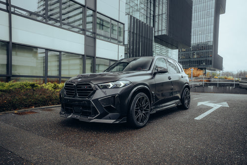 Black BMW X5M featuring a REVOZPORT carbon fiber body kit with aggressive front lip, side skirts, enlarged front air intakes, and sporty aero styling parked in a modern urban business district.