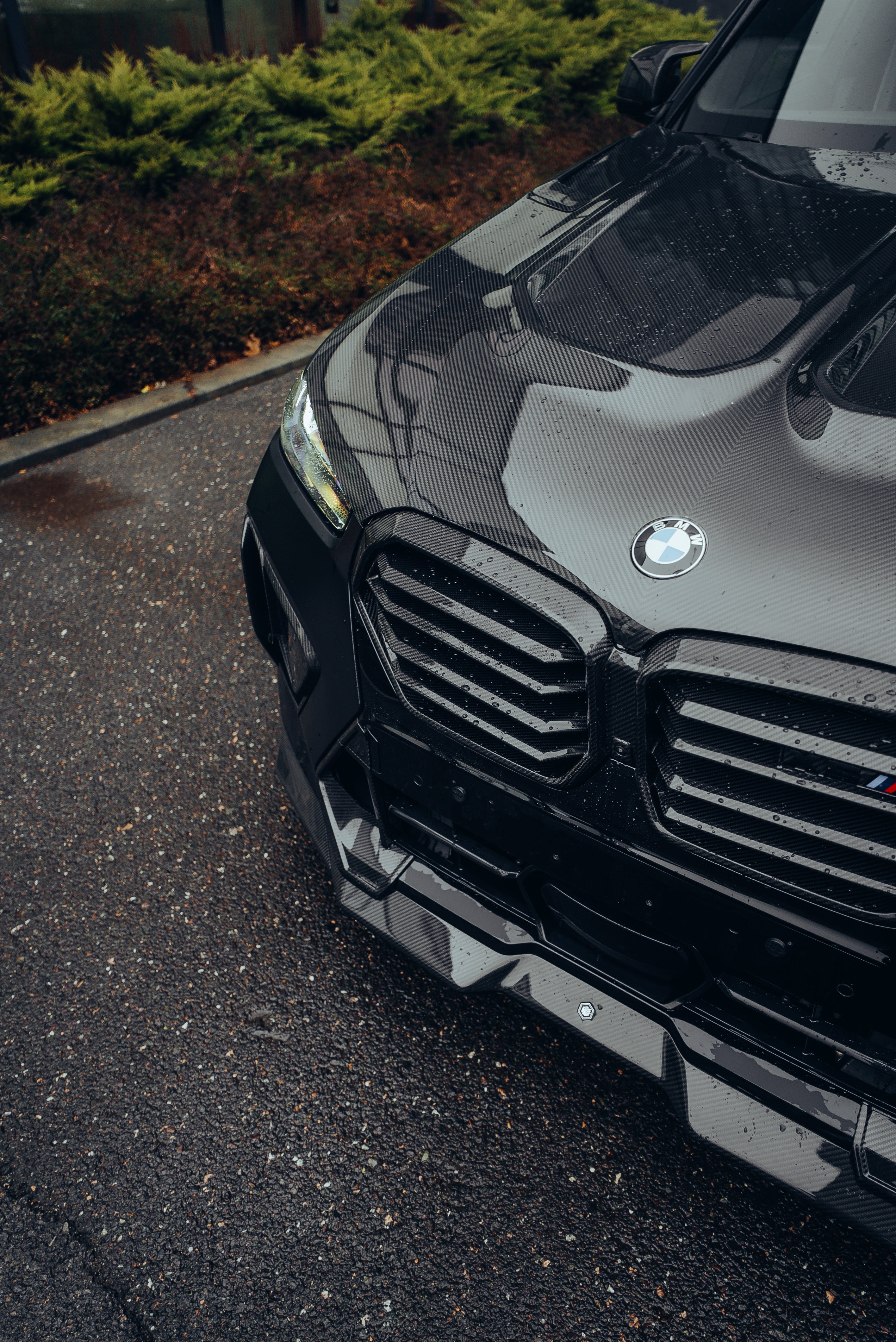Black BMW X5M featuring a REVOZPORT carbon fiber body kit with aggressive front lip, side skirts, enlarged front air intakes, and sporty aero styling parked in a modern urban business district.