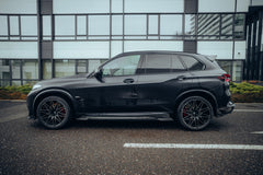 Black BMW X5M featuring a REVOZPORT carbon fiber body kit with aggressive front lip, side skirts, enlarged front air intakes, and sporty aero styling parked in a modern urban business district.