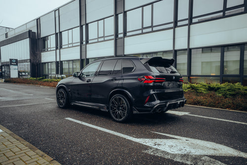 Black BMW X5M featuring a REVOZPORT carbon fiber body kit with aggressive front lip, side skirts, enlarged front air intakes, and sporty aero styling parked in a modern urban business district.