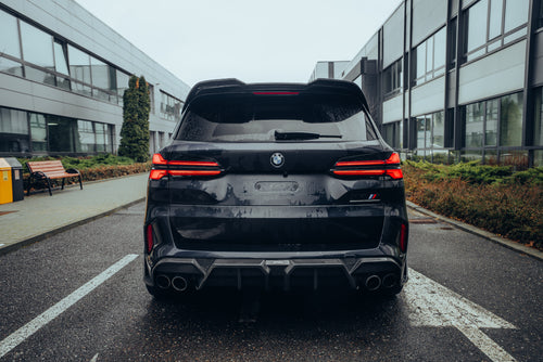 Black BMW X5M featuring a REVOZPORT carbon fiber body kit with aggressive front lip, side skirts, enlarged front air intakes, and sporty aero styling parked in a modern urban business district.
