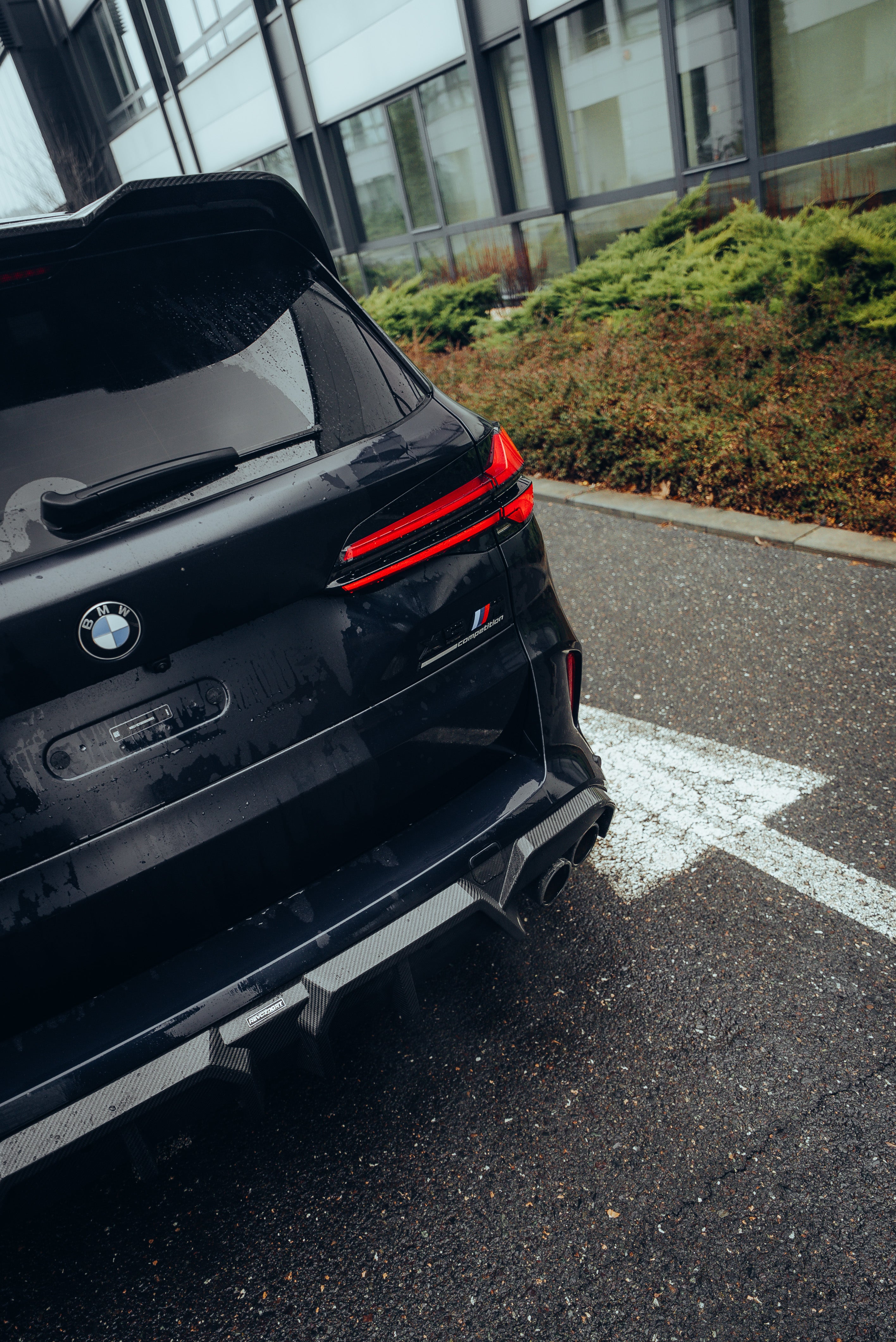 Black BMW X5M featuring a REVOZPORT carbon fiber body kit with aggressive front lip, side skirts, enlarged front air intakes, and sporty aero styling parked in a modern urban business district.