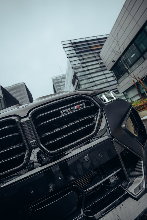 Black BMW X5M featuring a REVOZPORT carbon fiber body kit with aggressive front lip, side skirts, enlarged front air intakes, and sporty aero styling parked in a modern urban business district.