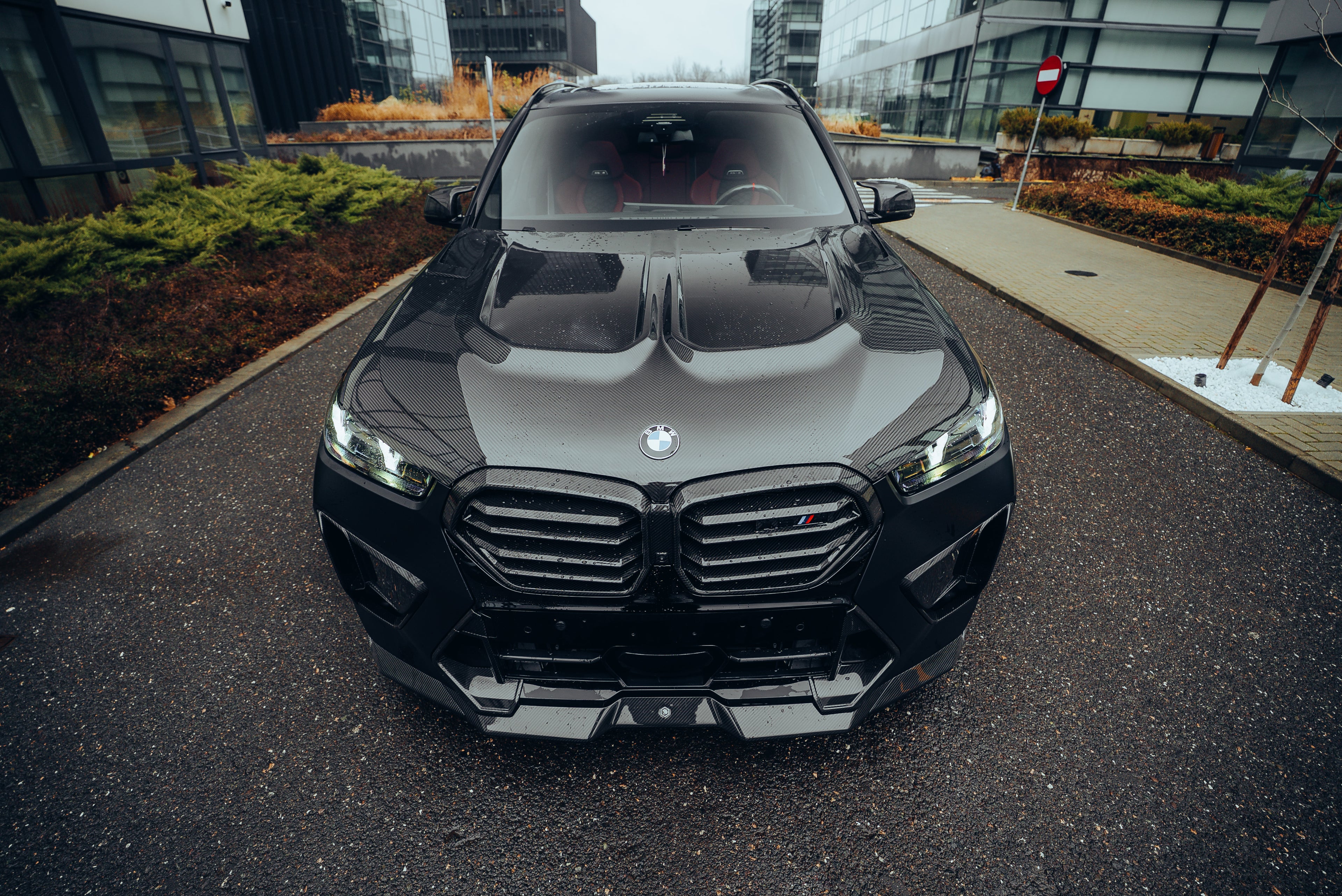 Black BMW X5M featuring a REVOZPORT carbon fiber body kit with aggressive front lip, side skirts, enlarged front air intakes, and sporty aero styling parked in a modern urban business district.