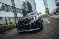 Black BMW X5M featuring a REVOZPORT carbon fiber body kit with aggressive front lip, side skirts, enlarged front air intakes, and sporty aero styling parked in a modern urban business district.
