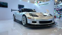 Close-up of a silver wide-body Chevrolet Corvette performance car displayed indoors, featuring aggressive aerodynamic bodywork, carbon fiber front splitter, vented hood, and lightweight multi-spoke wheels. The image highlights the custom motorsport styling and premium carbon fiber aero components under exhibition lighting.