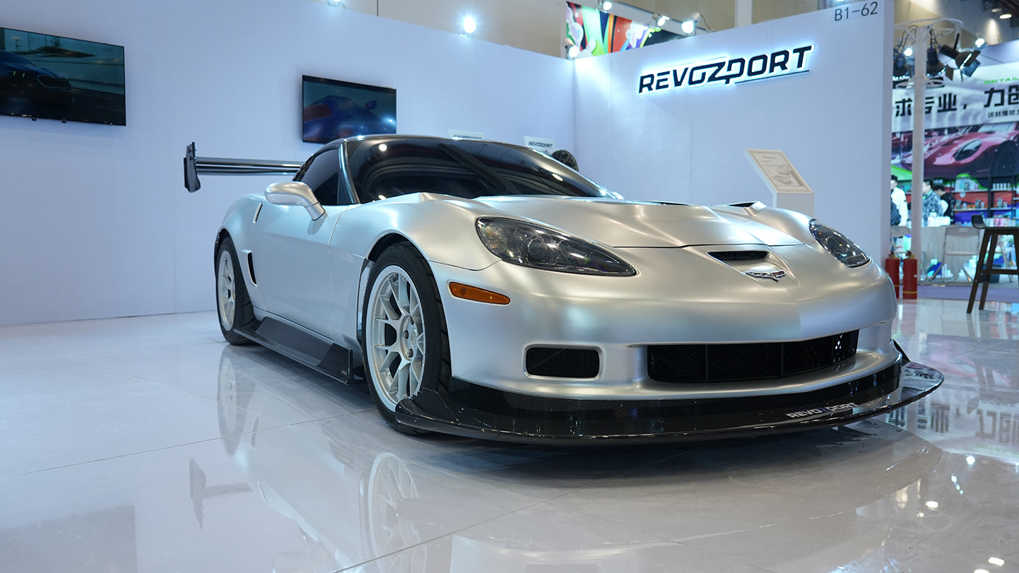 Close-up of a silver wide-body Chevrolet Corvette performance car displayed indoors, featuring aggressive aerodynamic bodywork, carbon fiber front splitter, vented hood, and lightweight multi-spoke wheels. The image highlights the custom motorsport styling and premium carbon fiber aero components under exhibition lighting.