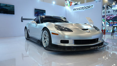 Close-up of a silver wide-body Chevrolet Corvette performance car displayed indoors, featuring aggressive aerodynamic bodywork, carbon fiber front splitter, vented hood, and lightweight multi-spoke wheels. The image highlights the custom motorsport styling and premium carbon fiber aero components under exhibition lighting.
