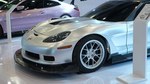 Close-up of a silver wide-body Chevrolet Corvette performance car displayed indoors, featuring aggressive aerodynamic bodywork, carbon fiber front splitter, vented hood, and lightweight multi-spoke wheels. The image highlights the custom motorsport styling and premium carbon fiber aero components under exhibition lighting.