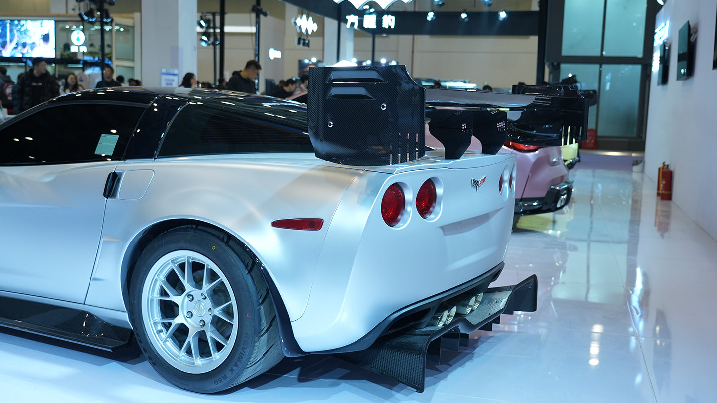Close-up of a silver wide-body Chevrolet Corvette performance car displayed indoors, featuring aggressive aerodynamic bodywork, carbon fiber front splitter, vented hood, and lightweight multi-spoke wheels. The image highlights the custom motorsport styling and premium carbon fiber aero components under exhibition lighting.