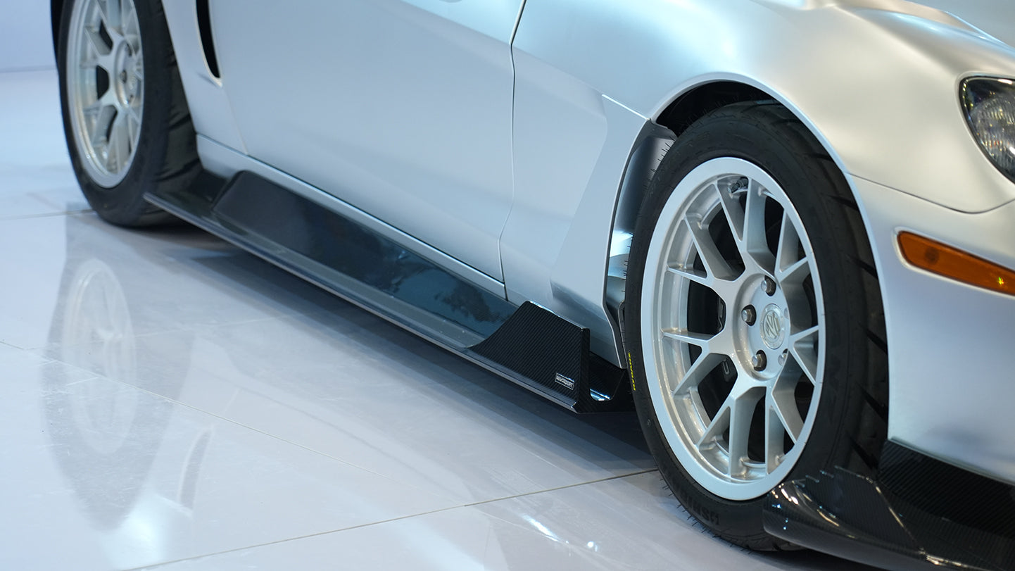 Close-up of a silver wide-body Chevrolet Corvette performance car displayed indoors, featuring aggressive aerodynamic bodywork, carbon fiber front splitter, vented hood, and lightweight multi-spoke wheels. The image highlights the custom motorsport styling and premium carbon fiber aero components under exhibition lighting.